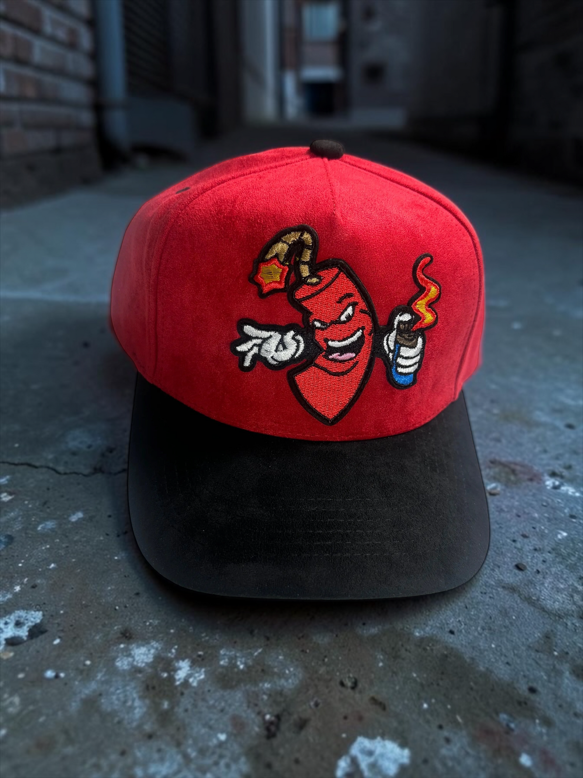 B-Is-TheBrand red suede baseball cap with a black brim and an embroidered mischievous firecracker character graphic.