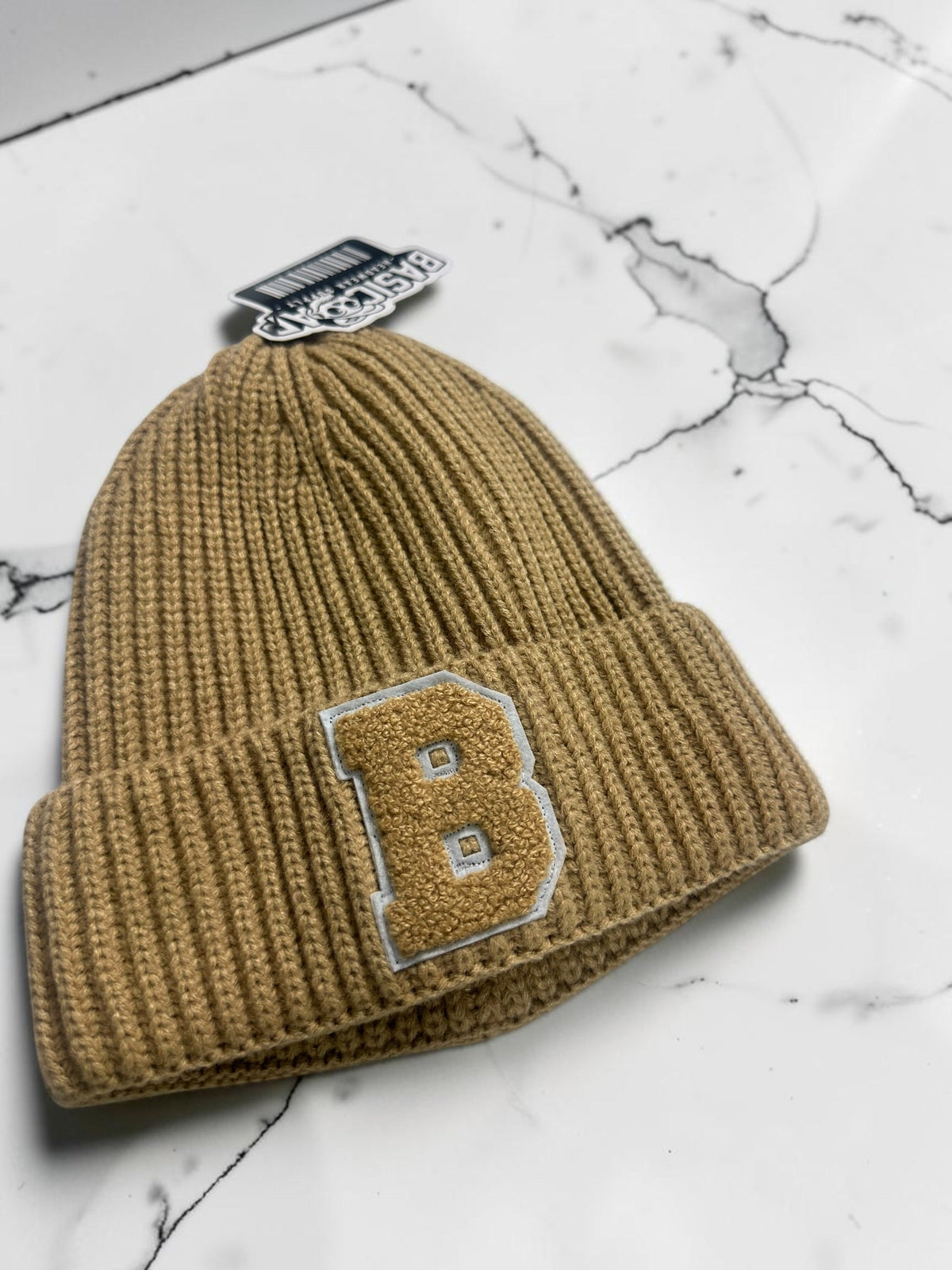 B-Is-TheBrand sand brown ribbed knit beanie with a textured collegiate B patch on a white marble surface